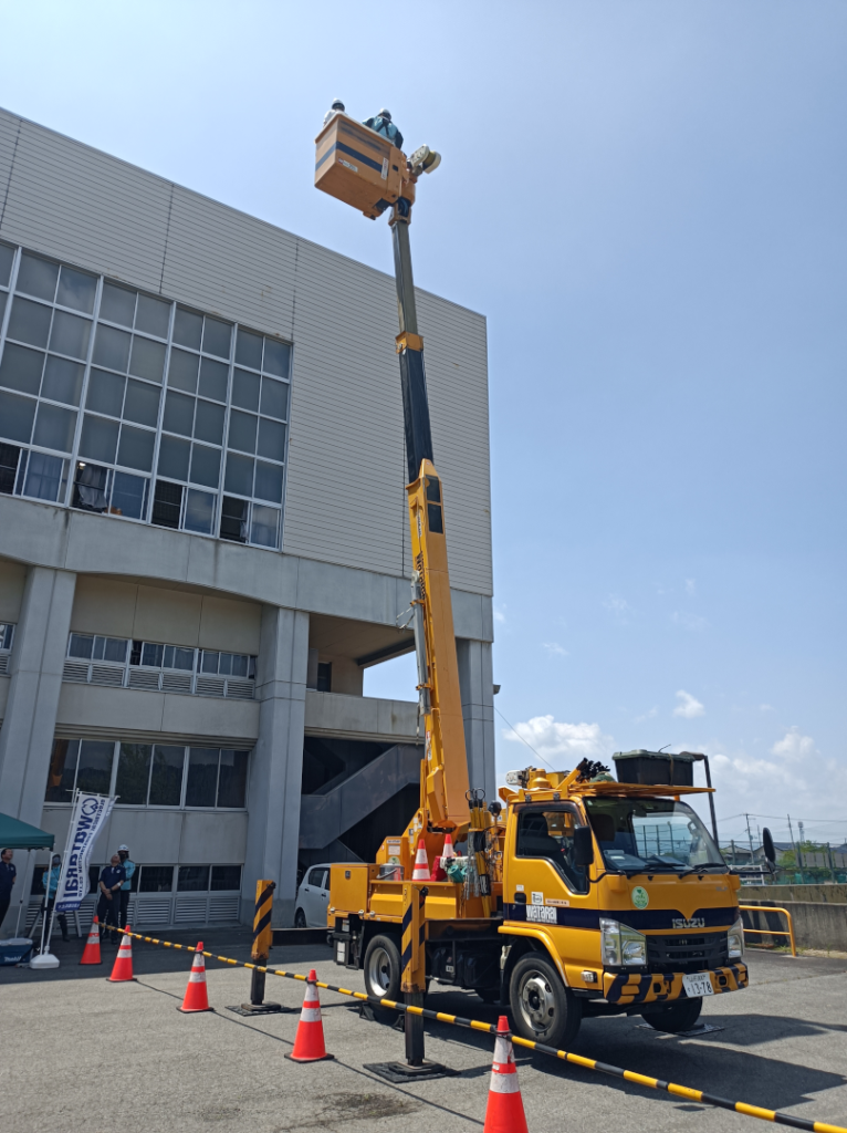 体育館前に黄色の高所作業車、ブームはほぼ鉛直に立ち上がり、体育館の天井付近ぐらいの高さまで伸びている。バケットには作業員と生徒さんが乗っている。