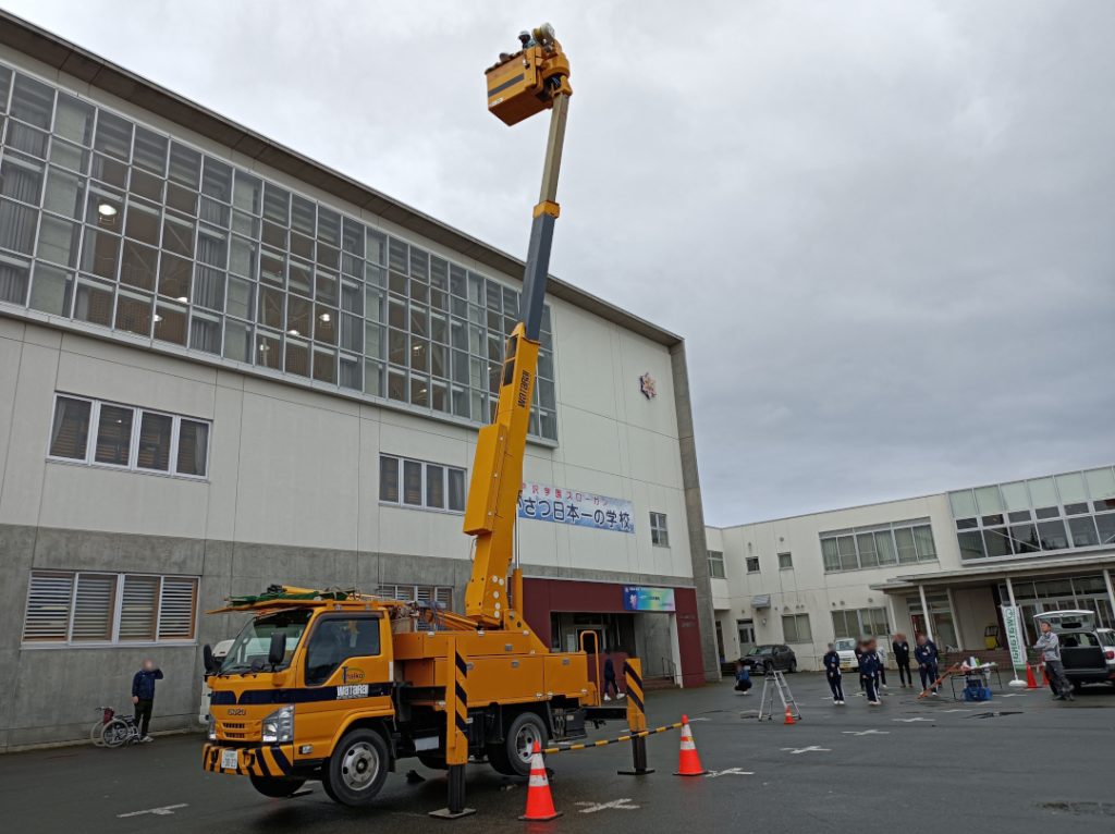 高所作業車に乗り、体育館ほどの高さまで上昇している写真